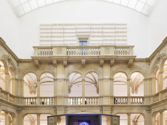 Das Bild zeigt einen Pavillon, der aus Spiegeln besteht, in einem historisch anmutendem Raum. Im Hintergrund sehen wir Bögen und Säulen. Auf dem Pavillon sehen wir im Spiegelbild ein Kunstwerk.