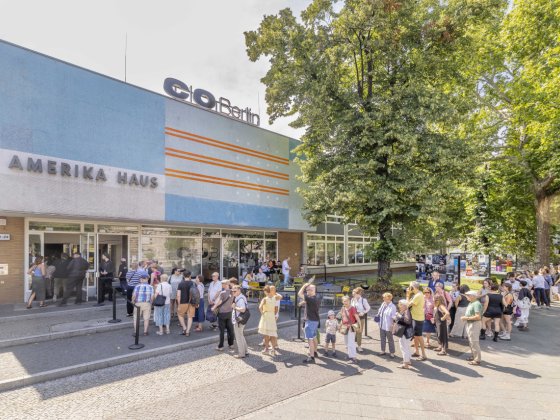 Außenansicht des C/O Berlin im Amerika Haus. Vor dem Eingang steht eine lange Schlange von Besucher:innen, daneben Sitzplätze unter Bäumen. Sonniger Tag, Plakate und Ausstellungsankündigungen an der Fassade.