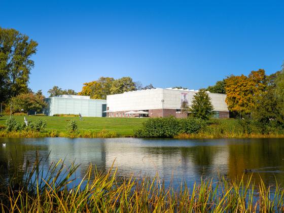 Die Kunsthalle Rostock und ihr neuer Anbau, das Schaudepot, betrachtet vom anderen Ufer des Schwanenteichs Die Gebäude sind links und rechts gerahmt von Bäumen. Vom Teich über eine Wiese mit leichter Steigung geht man auf die aufgespannten Schirme des Außenbereichs des Cafés in der Kunsthalle zu.