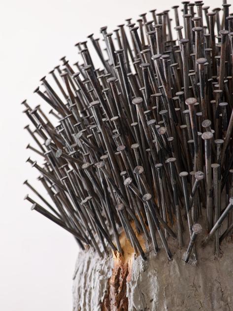 Zu sehen ist ein Ausschnitt und Nahsicht der Arbeit "Waldgarten" von Günther Uecker im Hochformat. Die Arbeit besteht aus einem Baumstamm, in den zig Nägel eingeschlagen wurden. Die Nägel sind nicht komplett im Stamm versenkt, sodass eine Art Kuppel, Stachel-Frisur oder abstrakte Baumkrone ensteht. 