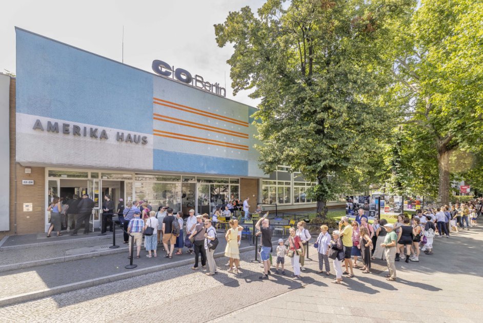 Außenansicht des C/O Berlin im Amerika Haus. Vor dem Eingang steht eine lange Schlange von Besucher:innen, daneben Sitzplätze unter Bäumen. Sonniger Tag, Plakate und Ausstellungsankündigungen an der Fassade.