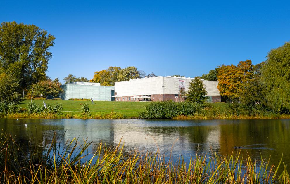 Die Kunsthalle Rostock und ihr neuer Anbau, das Schaudepot, betrachtet vom anderen Ufer des Schwanenteichs Die Gebäude sind links und rechts gerahmt von Bäumen. Vom Teich über eine Wiese mit leichter Steigung geht man auf die aufgespannten Schirme des Außenbereichs des Cafés in der Kunsthalle zu.