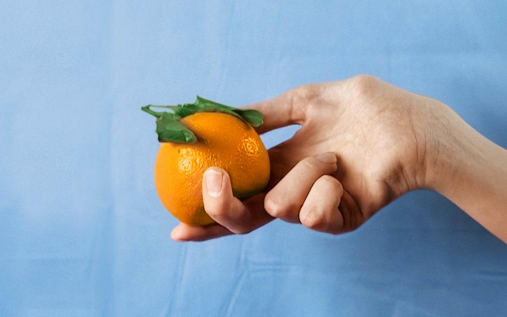 A woman's hand holds a bright orange against a light blue, slightly crumpled background of fabric.