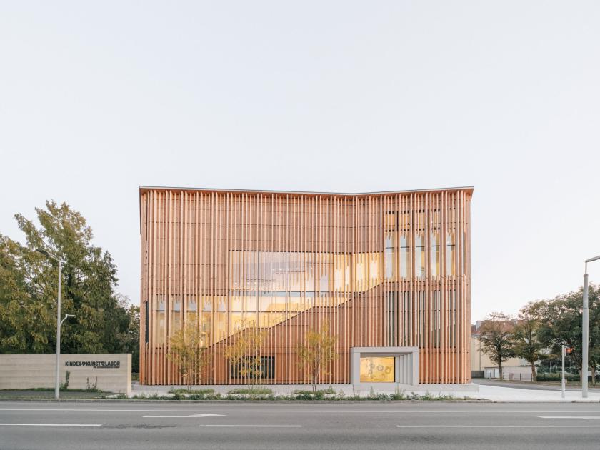Zu sehen ist das KinderKunstLabor für zeitgenössische Kunst in der Frontalansicht: Die Fassade des Gebäudes besteht aus Holz, Teile des Innenbereichs sind beleuchtet, links und rechts sind Bäume zu sehen. 