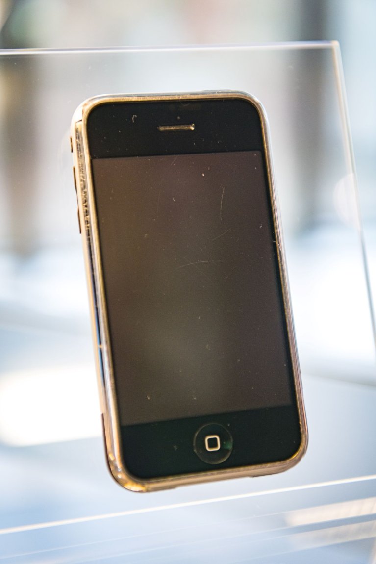 Smartphone mit Homebutton auf transparentem Halter ausgestellt; schwarzes Display, Silberrahmen.