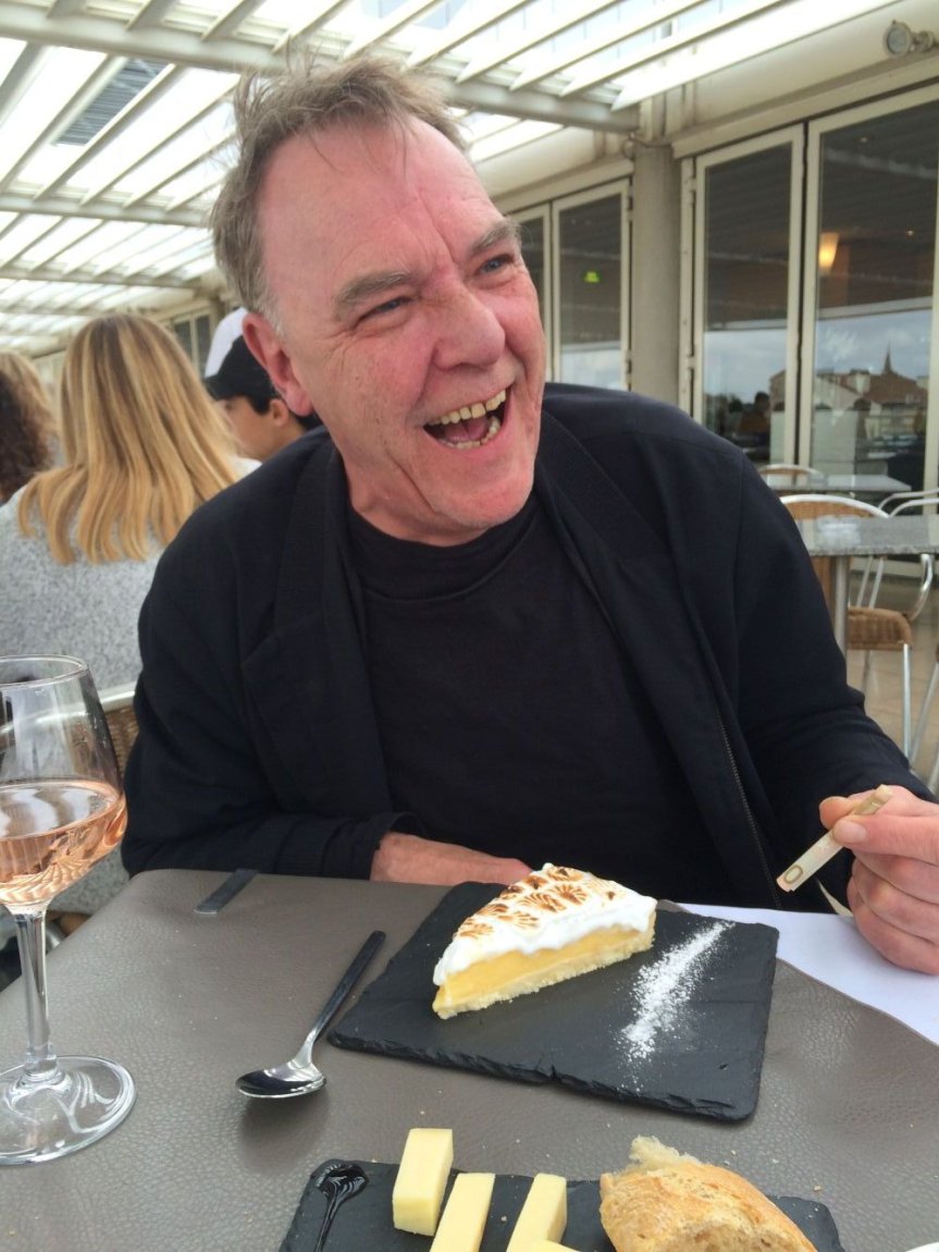 Porträtaufnahme: lachende Person auf Caféterrasse, Dessert, Käse, Rosé, Zigarette vor sich.