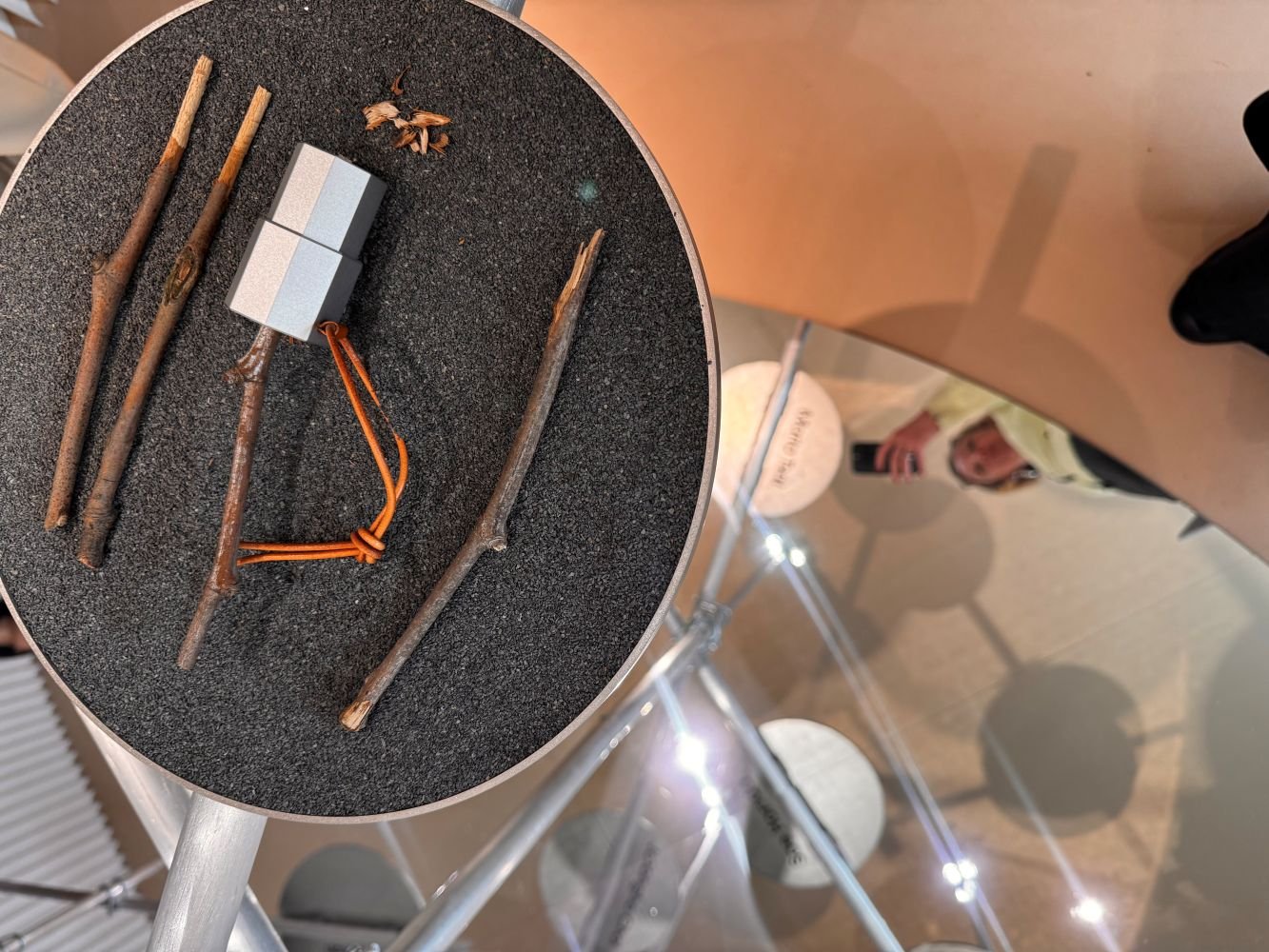 Foto: Kleine Assemblage aus Zweigen, weißem Quader und orangen Schnüren auf runder, grauer Platte.