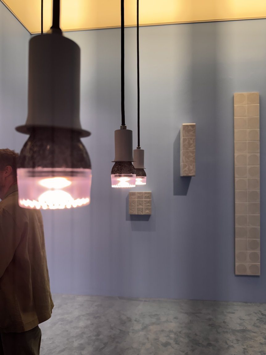 Installationsansicht: Vorne drei Pendelleuchten; blaue Wand mit rechteckigen Reliefs; Person links