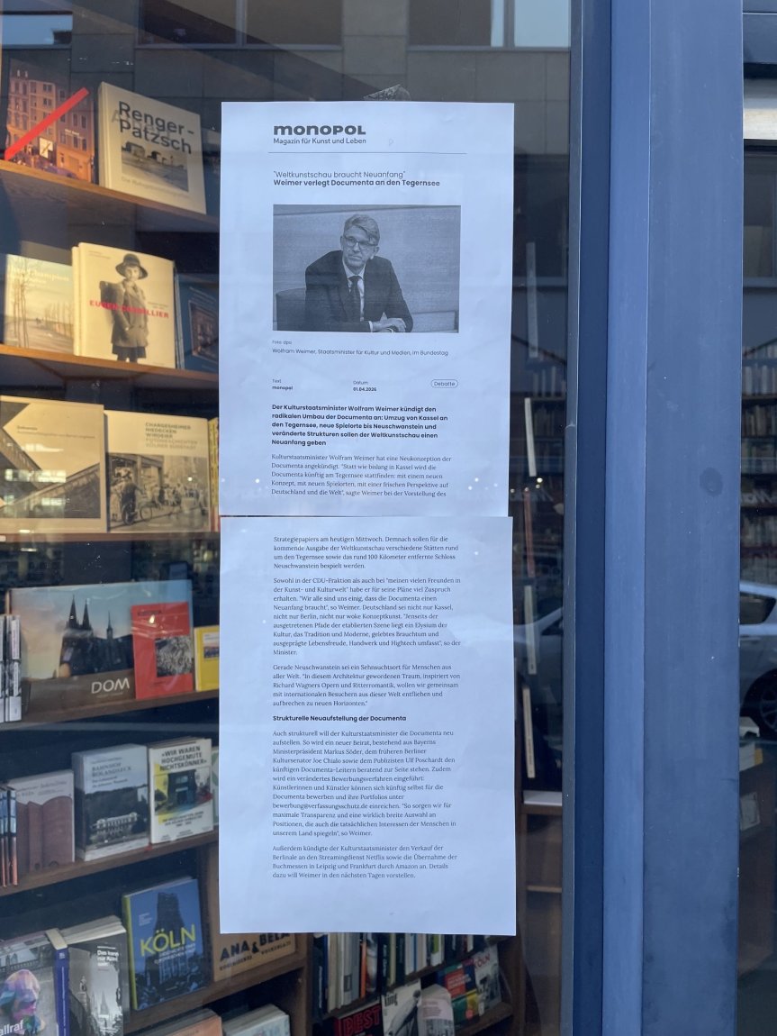 Zweiseitiger Aushang mit Text und s/w-Foto am Buchladenfenster; dahinter Bücherregale.