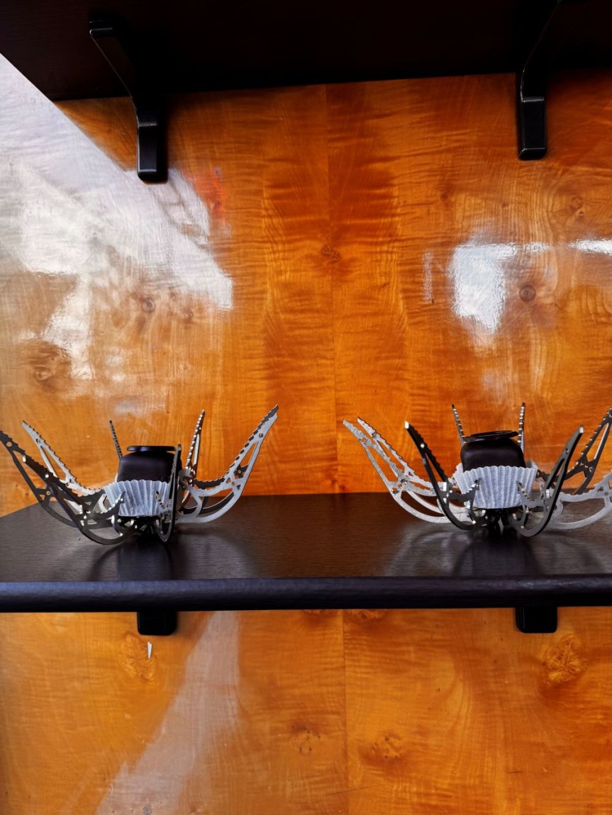 Zwei kleine spinnenartige Skulpturen aus Drähten und Bauteilen auf schwarzem Regal vor Holzwand.