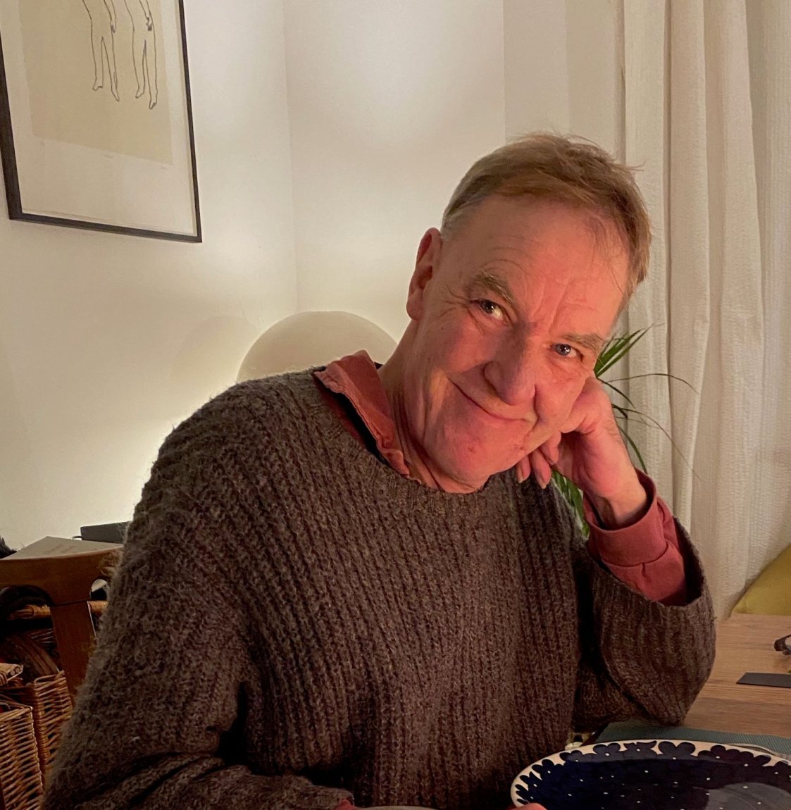 Foto: Porträt einer Person am Esstisch, Hand am Kinn, Blick zur Kamera, warmes Licht, Teller vorn.
