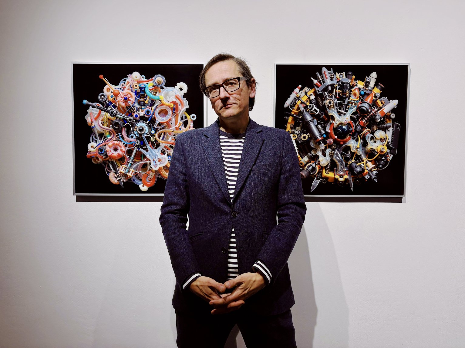 Foto: Person in Galerie vor zwei gerahmten, kreisförmigen Bildern; stehend, Hände gefaltet.