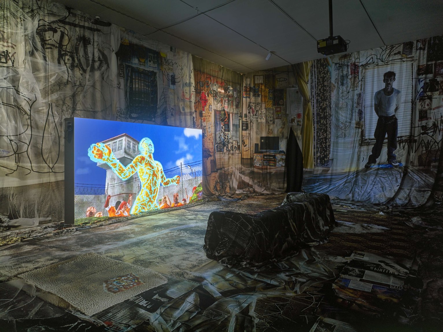 Installationsansicht: Videoinstallation, großer Screen links; Sitzbank; Wände mit Collagen; Beamer.