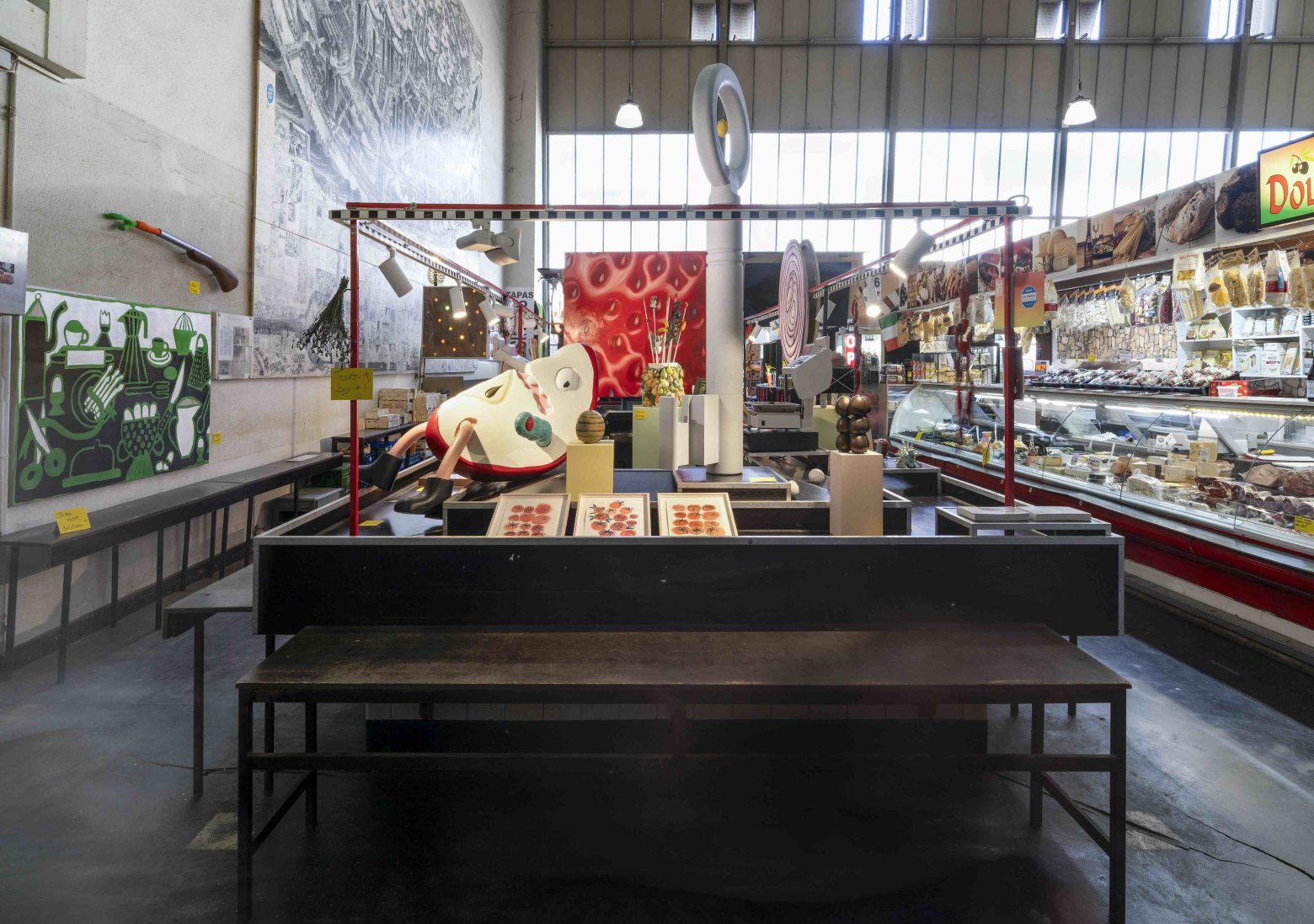 Installationsansicht in Markthalle: Gemälde, Drucke und Objekte auf Marktstand, umgeben von Theken.