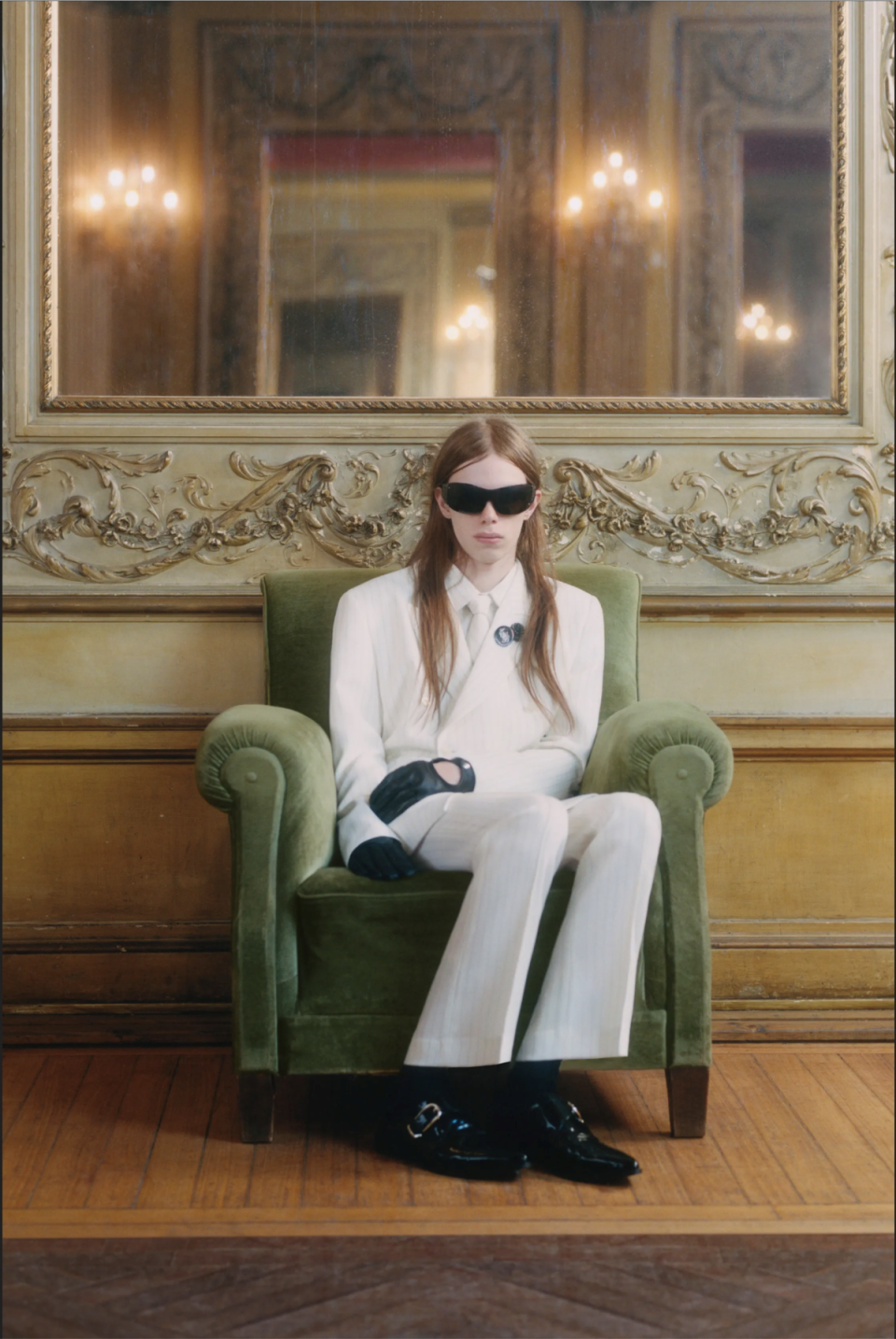 Fotografie: Person im weißen Anzug mit Sonnenbrille im grünen Sessel, vergoldeter Spiegel dahinter.