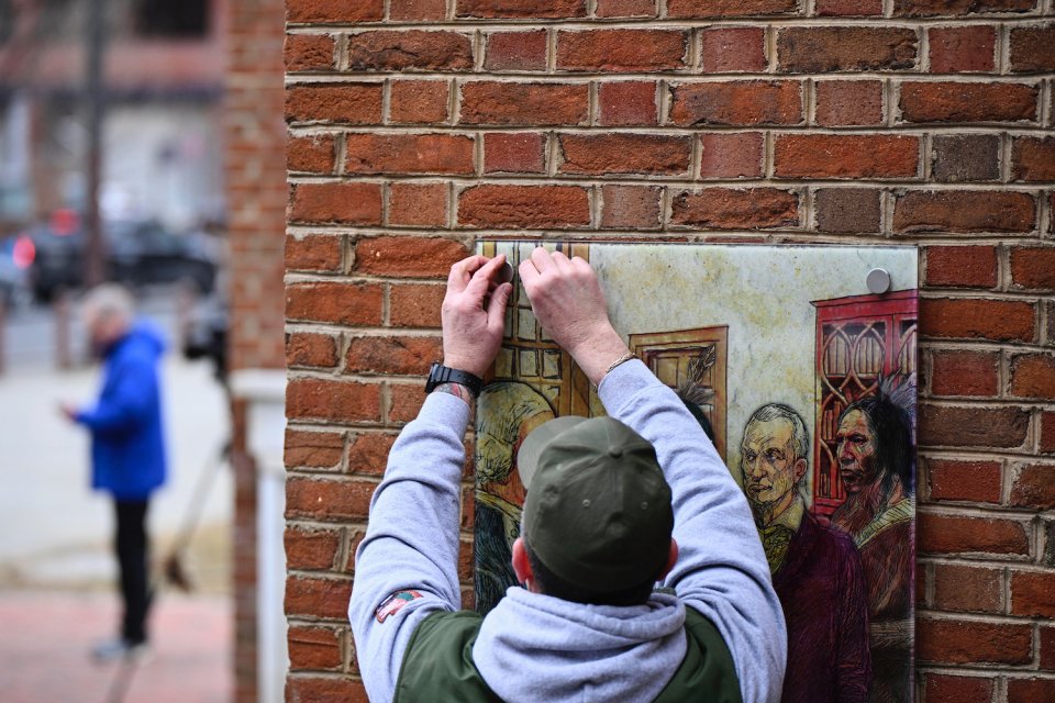 Foto: Techniker montiert Bildtafel mit Porträts an Backsteinwand; Außeninstallation.