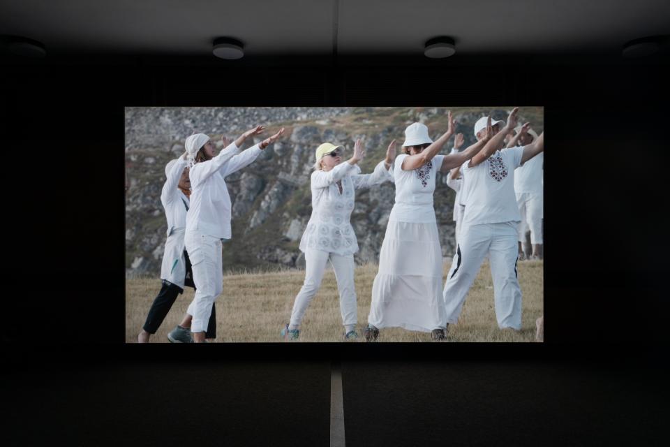 Installationsansicht: großformatige Videoprojektion in dunklem Raum; Gruppe in Weiß hebt Arme.