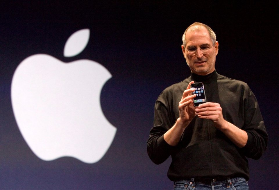 Foto: Produktpräsentation auf Bühne; Person hält Smartphone, großes Apple-Logo im Hintergrund.