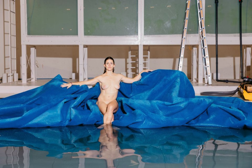 Fotografie: Nackte Person auf blauer Plane am Hallenbadbecken, Leiter im Hintergrund, Spiegelung.