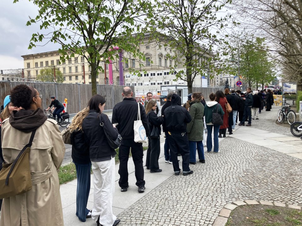 Lange Schlange für eine Kunstausstellung entlang eines baumbestandenen Gehwegs im Freien.