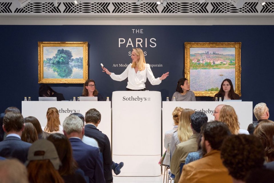 Fotografie: Sotheby’s Auktion; Auktionatorin am Pult, Publikum, zwei Gemälde an blauer Wand.