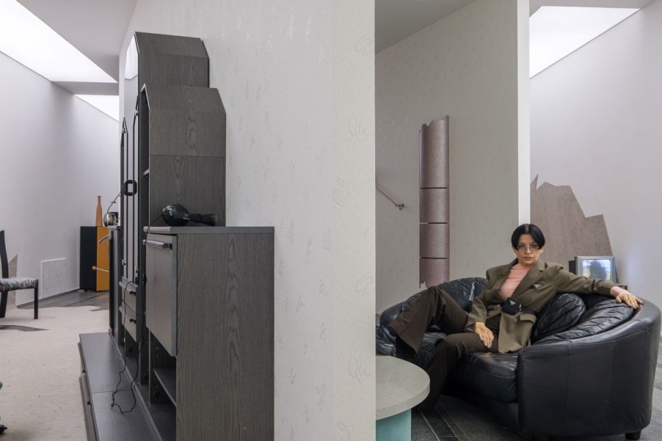 Installationsansicht mit Trennwand; links Möbelensemble, rechts Person auf schwarzem Sofa.