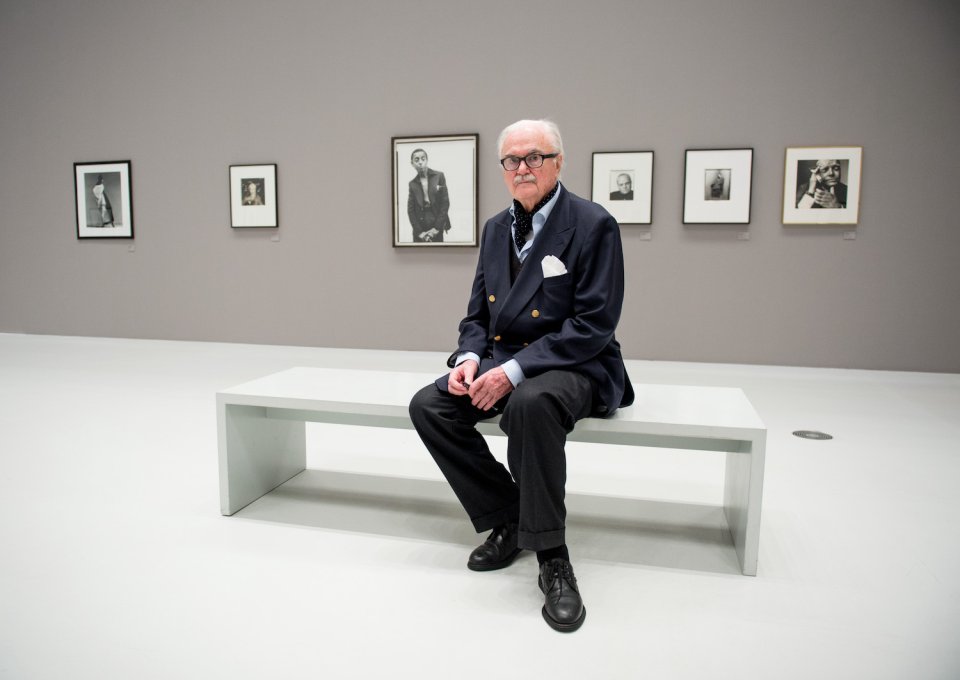 Porträtfoto: älterer Mann auf Bank im Ausstellungsraum; Reihe s/w‑Fotografien an grauer Wand.