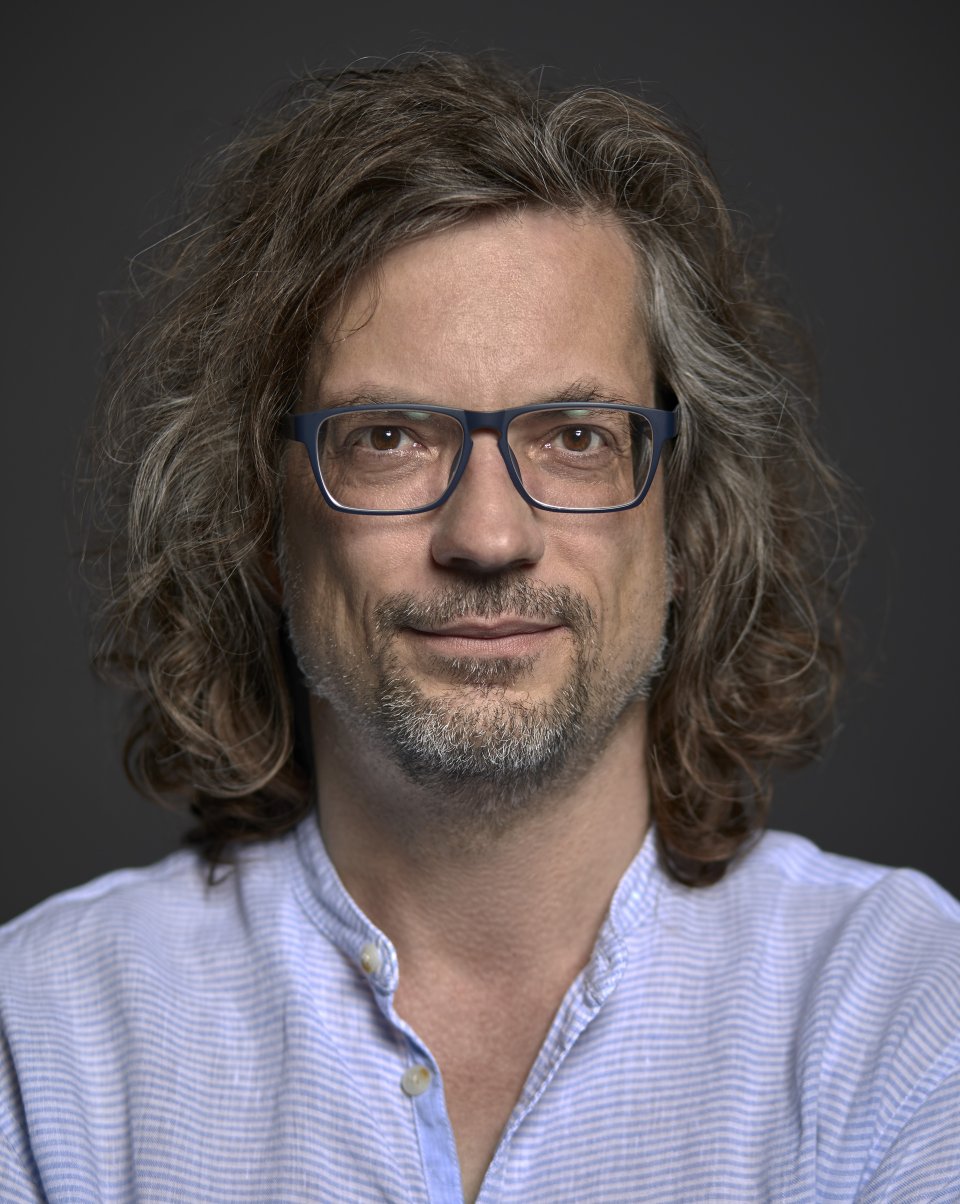 Porträtfoto: Person mit schulterlangem Haar und blauer Brille, neutraler Blick, Studiohintergrund.