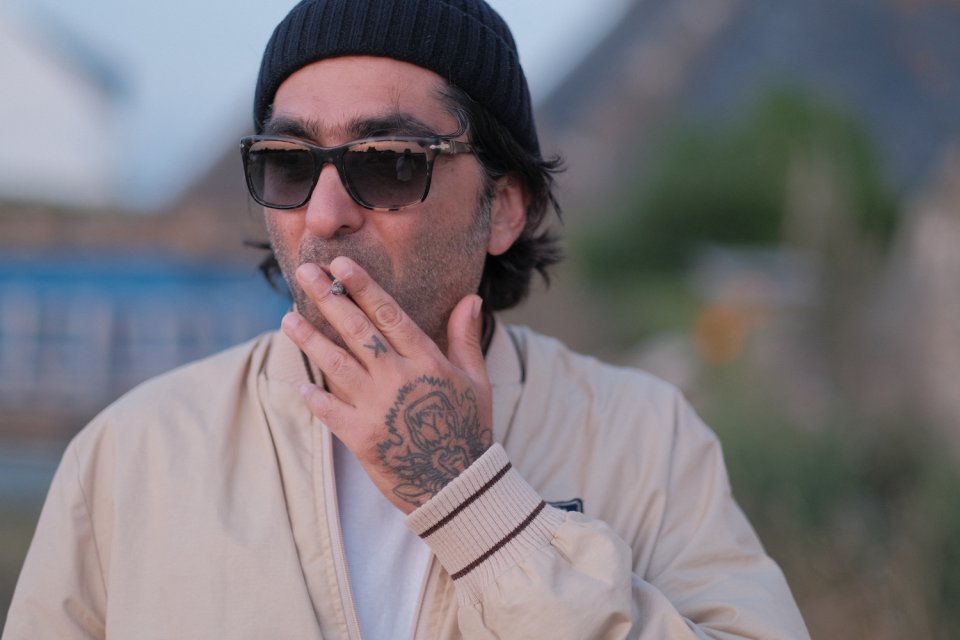 Porträtfoto einer Person im Freien, mit Mütze und Sonnenbrille, rauchend, Handtattoo, beiger Jacke.