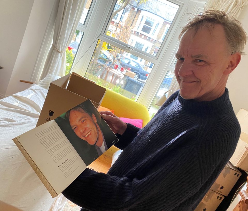 Mann in hellem Zimmer hält offenes Buch mit Porträtfoto und Text, lächelt zur Kamera.