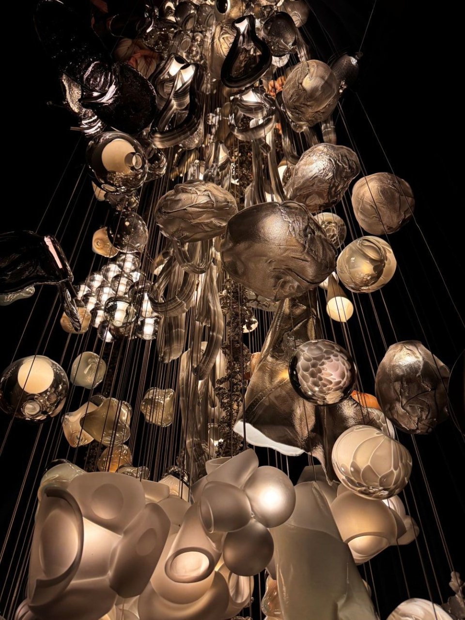 Hängende Lichtinstallation aus zahlreichen Glasformen, dichte Cluster in dunklem Raum, Untersicht.