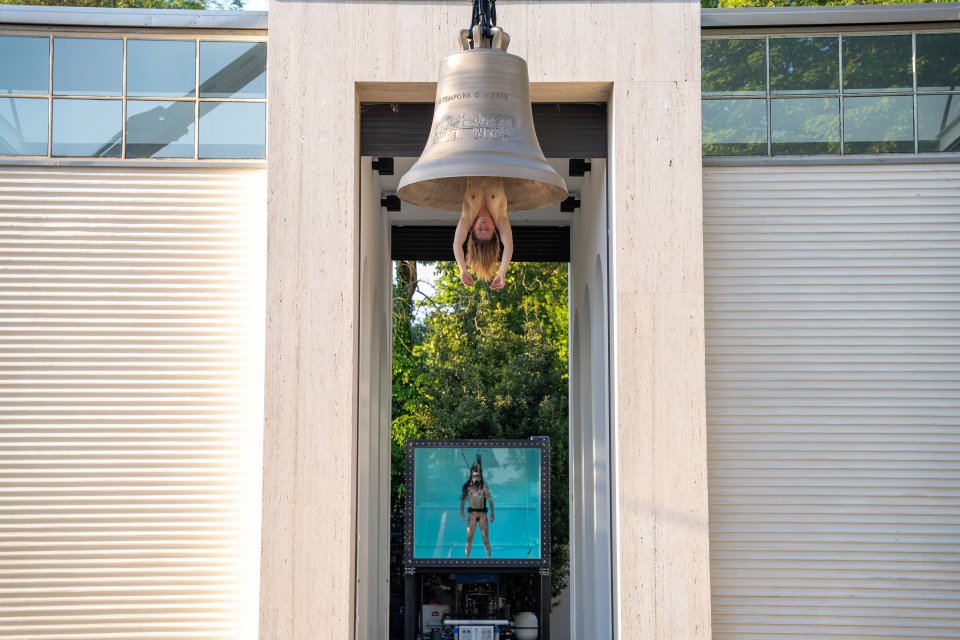 Installationsansicht am Pavillonportal: große hängende Glocke mit Person; unten Video.