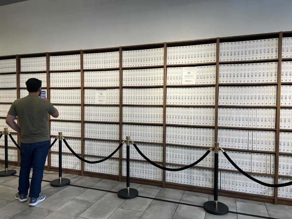 Installationsansicht: wandfüllende Holzregale mit Reihen identischer weißer Bücher; Besucher davor.