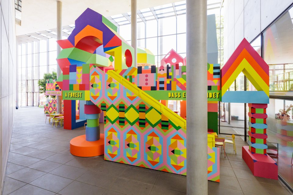 Installationsansicht: raumfüllende, mehrstöckige, bunte geometrische Struktur mit Treppe in Halle
