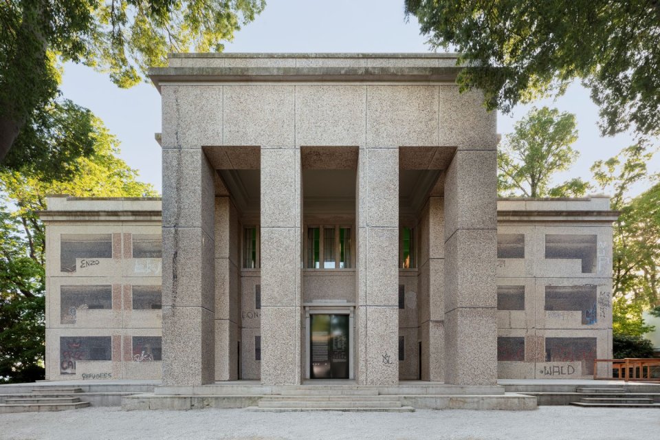 Deutscher Pavillon, Außenansicht; hohe Pfeiler, symmetrische Steinfassade, Bäume im Hintergrund.