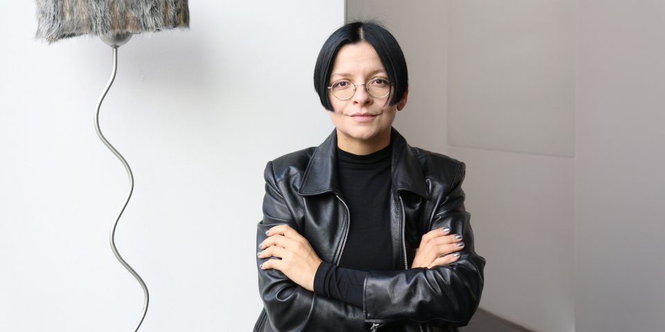 Fotoporträt einer Person mit verschränkten Armen in schwarzer Lederjacke, neben skulpturaler Lampe.