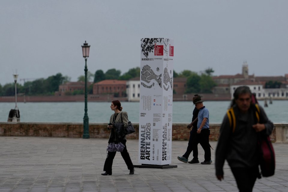 Fotografie: Stele der Kunstbiennale am Ufer; Passanten, Lagune, Laterne im Hintergrund.