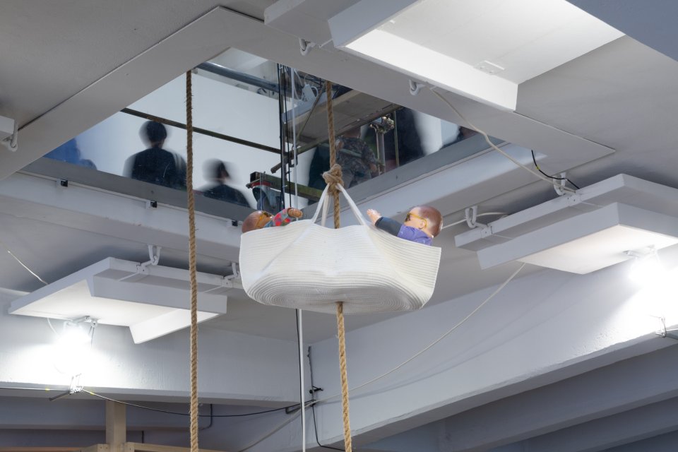 Installationsansicht: Person in weißem Hängesack an Seilen unter Spiegeldecke mit Reflexionen.