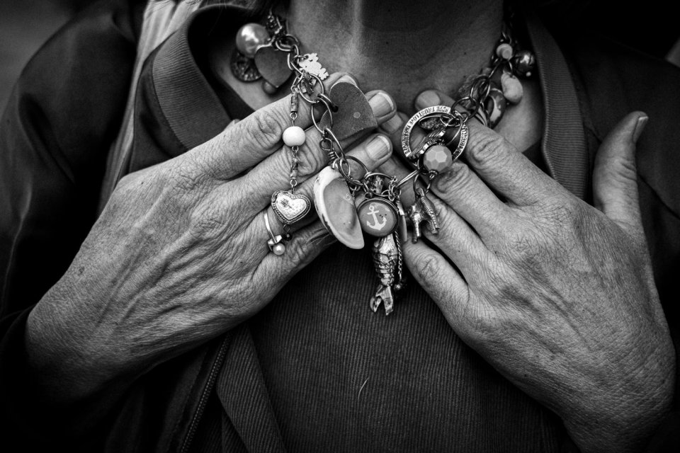 Schwarzweiß-Foto: faltige Hände berühren Kette mit zahlreichen Anhängern am Hals, Nahaufnahme.