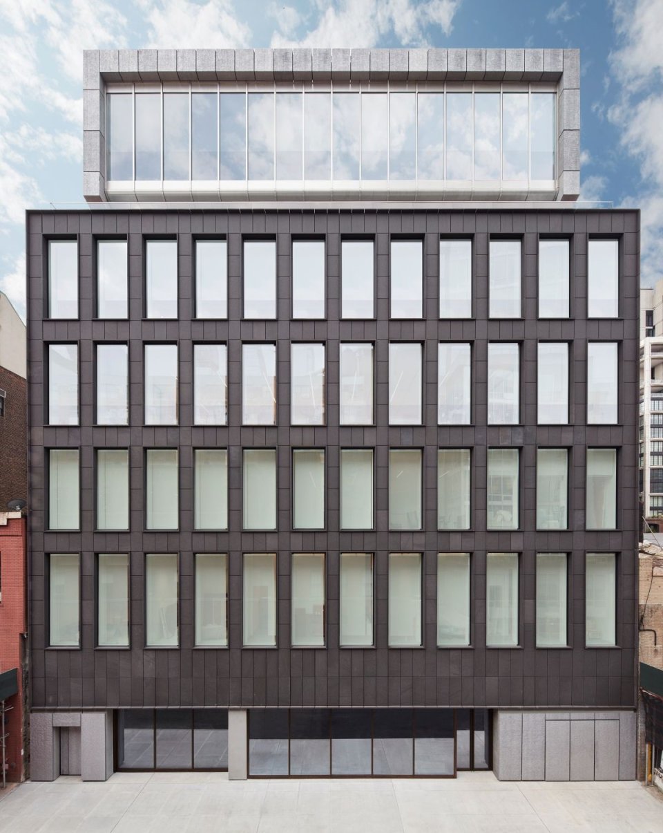 Außenansicht einer sechsgeschossigen Galerie: dunkle Rasterfassade, gläserner Dachaufbau.
