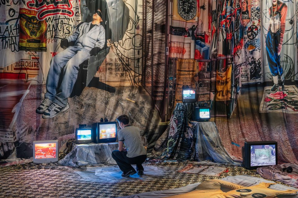 Installationsansicht: mehrere CRT-Monitore am Boden vor großformatigen Textilprints; Besucher kniet.
