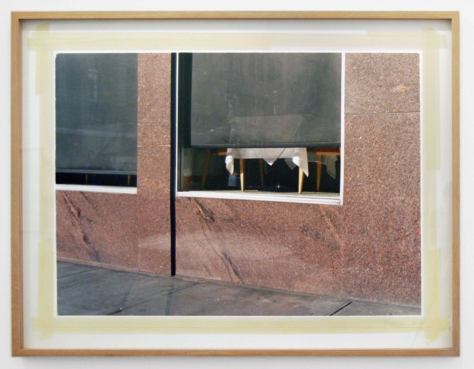 Gerahmte Fotografie: Schaufensterfassade aus rötlichem Stein, zerrissenes Papier im Fenster.