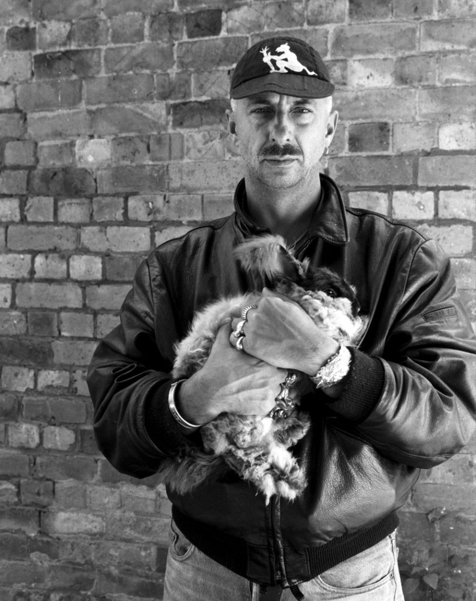 Schwarzweißfoto: Person mit Basecap und Lederjacke hält Katze vor Backsteinwand, frontal.