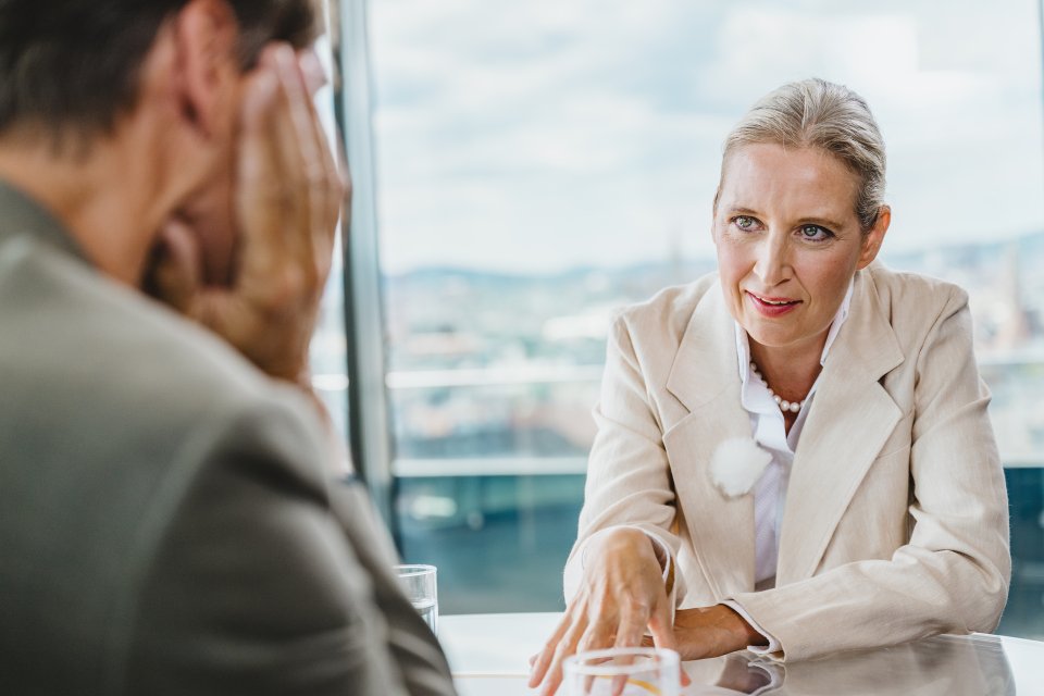 Foto: Interview am Tisch, Frau spricht und gestikuliert; Mann im Vordergrund unscharf, Stadtblick.