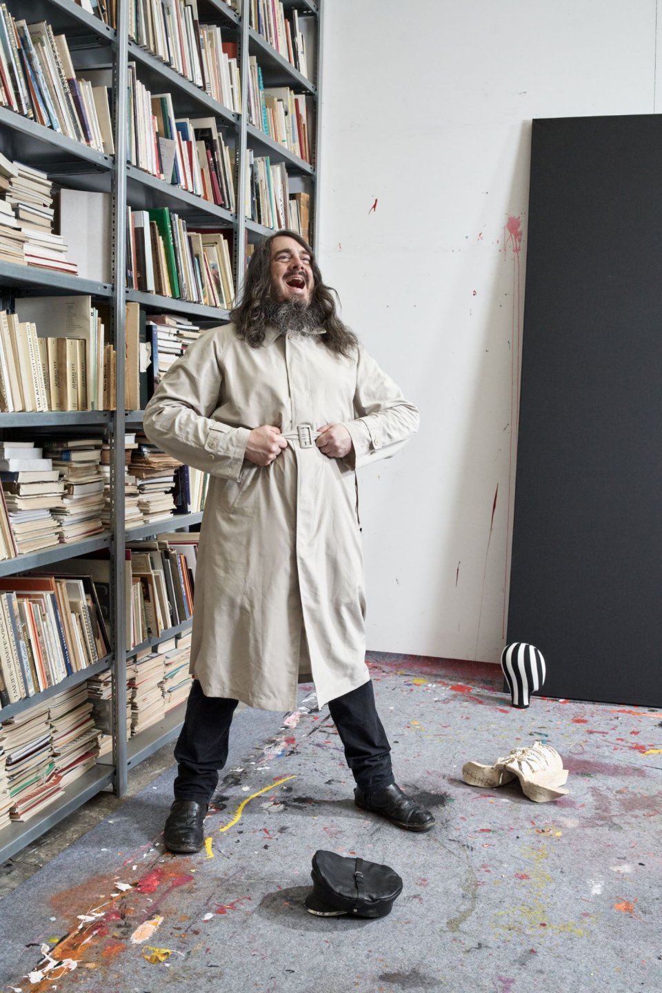 Porträtfoto Jonathan Meese: Person im Atelier vor Bücherregalen; Mantel offen; schwarzes Panel, Objekte am Boden.