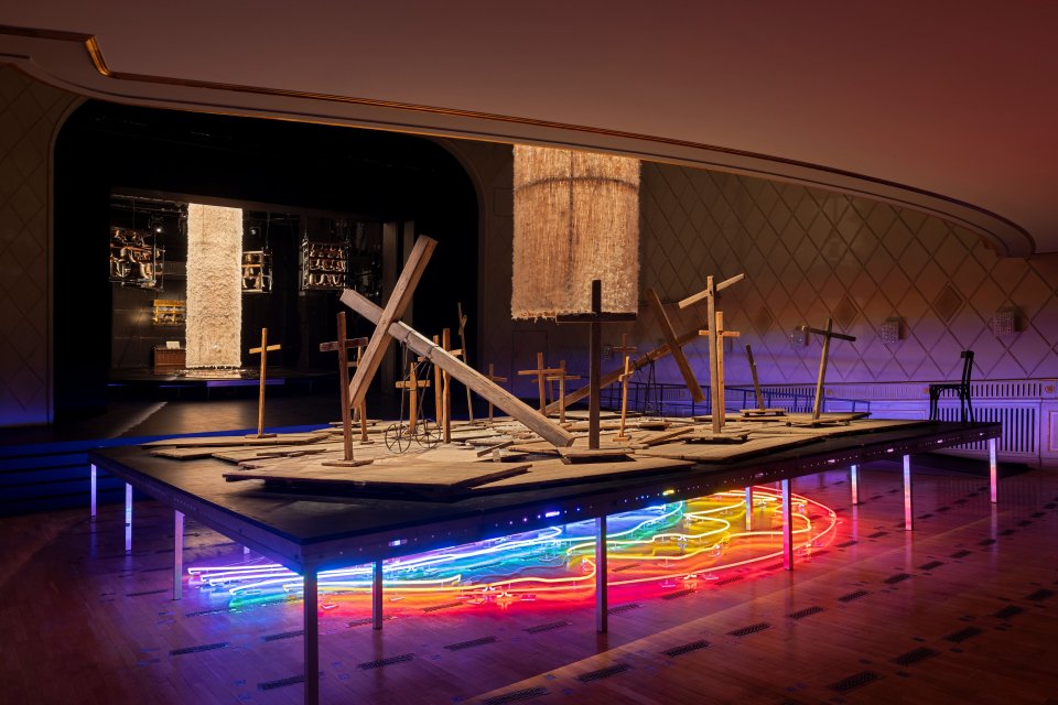 Installationsansicht: Holzkreuze auf Podest; darunter wellenförmiges Neon in Regenbogenfarben.