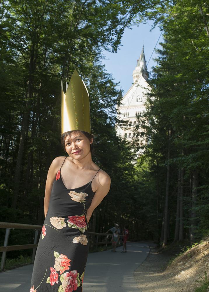 Was macht die Thailänderin vor dem Schloss Neuschwanstein? Sicher ist: Sie ist hier nicht allein. Touristen aus aller Welt lieben diesen gebauten Kindertraum. Auch die Künstlerin Orawan Arunrak hat schon als Kind von Neuschwanstein geträumt – eine nachgebaute Version stand in ihrer Heimat Bangkok. Und als sie endlich das echte Neuschwanstein besuchte, hatte sie das nötige Accessoire dabei und krönte sich selbst zur Königin.