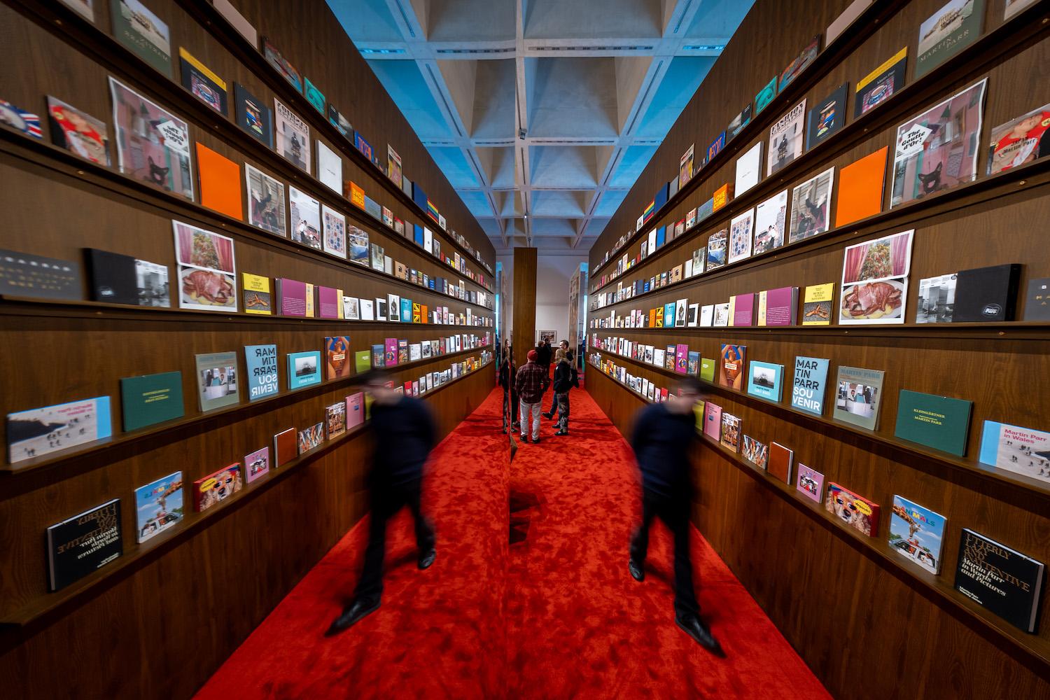 Eine Wand mit sämtlichen bislang veröffentlichten Fotobüchern des britischen Magnum-Fotografen Martin Parr spiegelt sich in der „Lounge“ der Ausstellung „Grand Hotel Parr – Fotobücher von Martin Parr“ im Neuen Museum Nürnberg