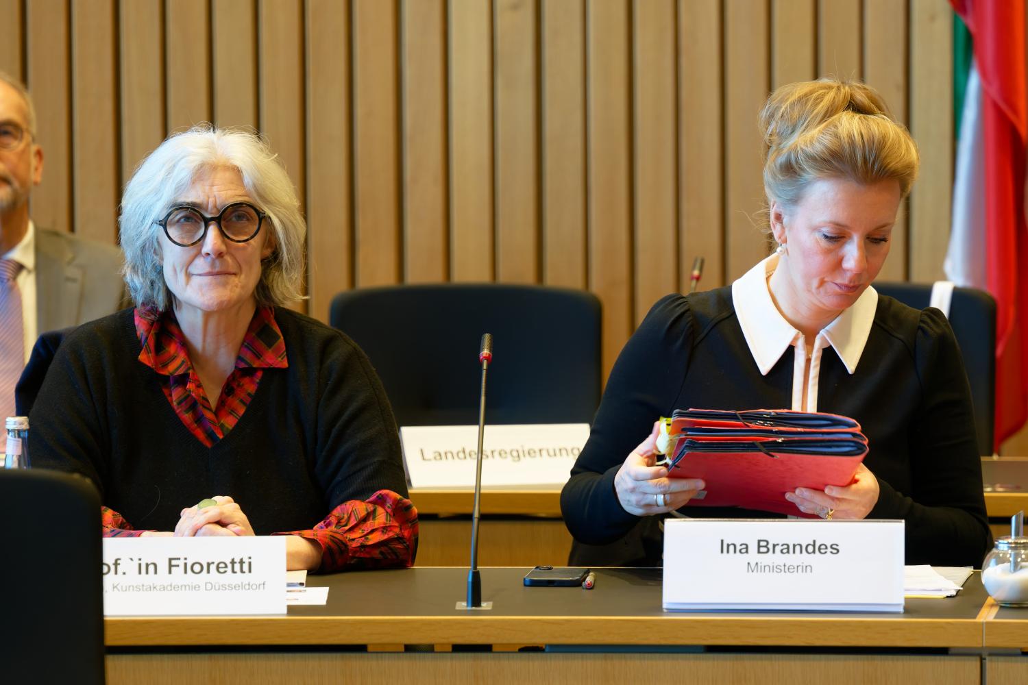 Foto: Zwei Frauen bei einer Ausschusssitzung, Mikrofone und Akten, Holzvertäfelung, Flagge