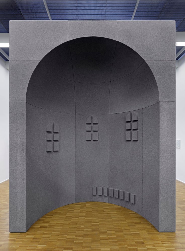 Installationsansicht einer raumhohen grauen Wandskulptur mit Rundbogennische und Kreuzfenstern.